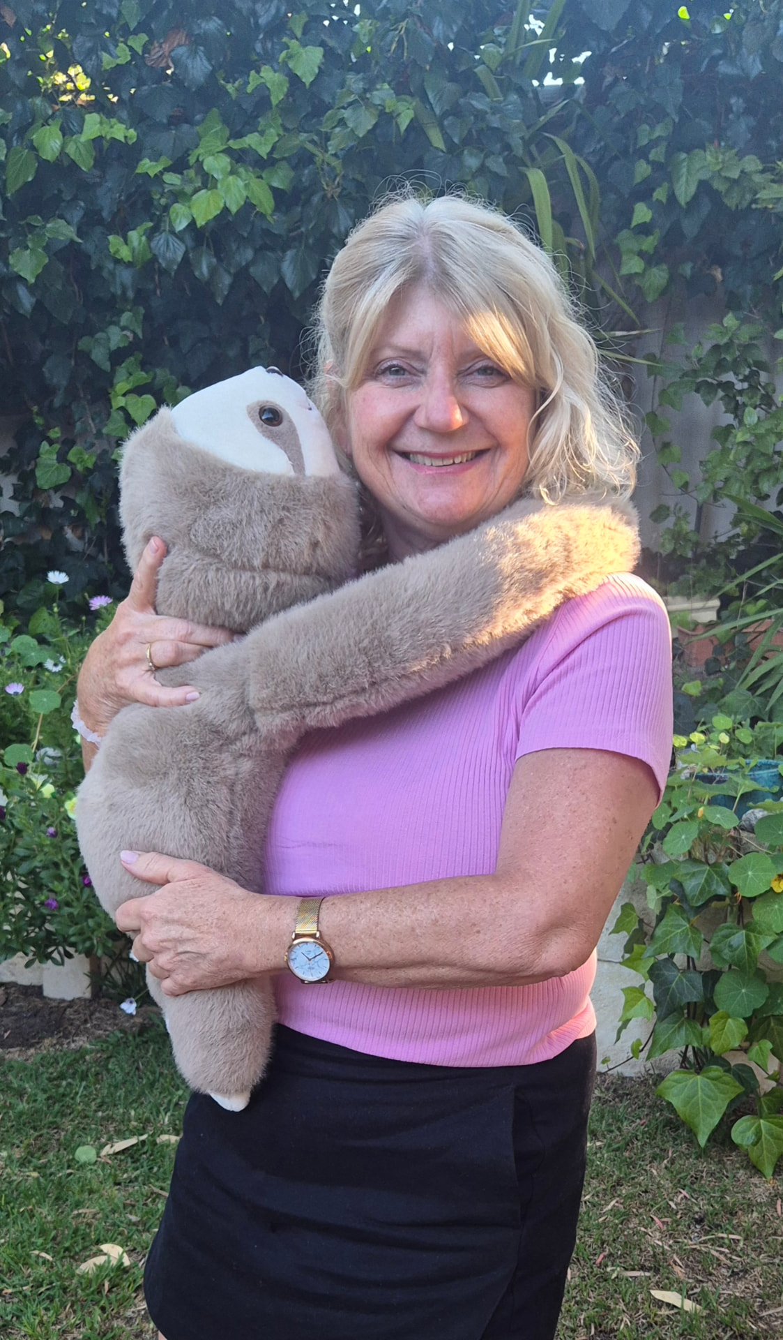 Carolyn smiling outdoors with the weighted sloth Sally, its long arms draped over her shoulders